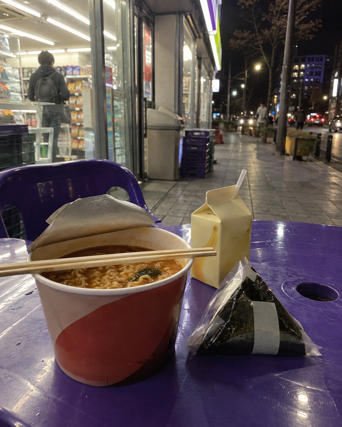 夜のコンビニの外のテーブルに、カップラーメンとおにぎりとバナナ牛乳
